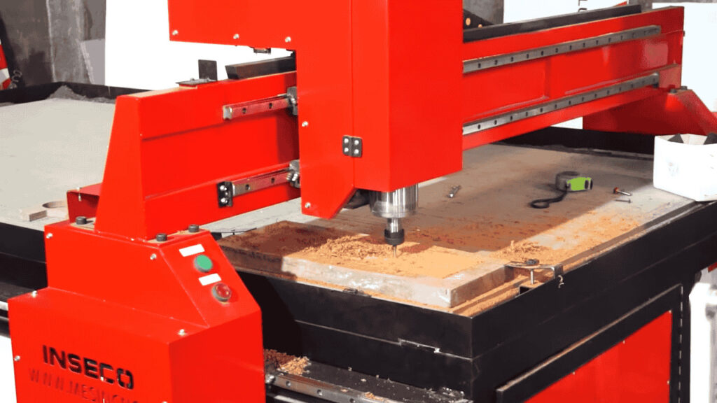 Keunggulan Mesin CNC Router / Mesin Ukir Potong Kayu 4 Mesin CNC Router merk INSECO berwarna merah sedang melakukan proses pengukiran pada kayu. Terlihat spindle mesin sedang bekerja dengan serbuk kayu berserakan di permukaan bahan menunjukkan proses pengerjaan